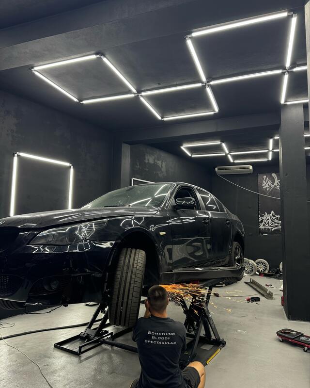 Czarne auto na stojakach pod sufitem z sześciokątnymi lampami LED. Mechanik, widoczny od tyłu, szlifuje, lecą iskry.